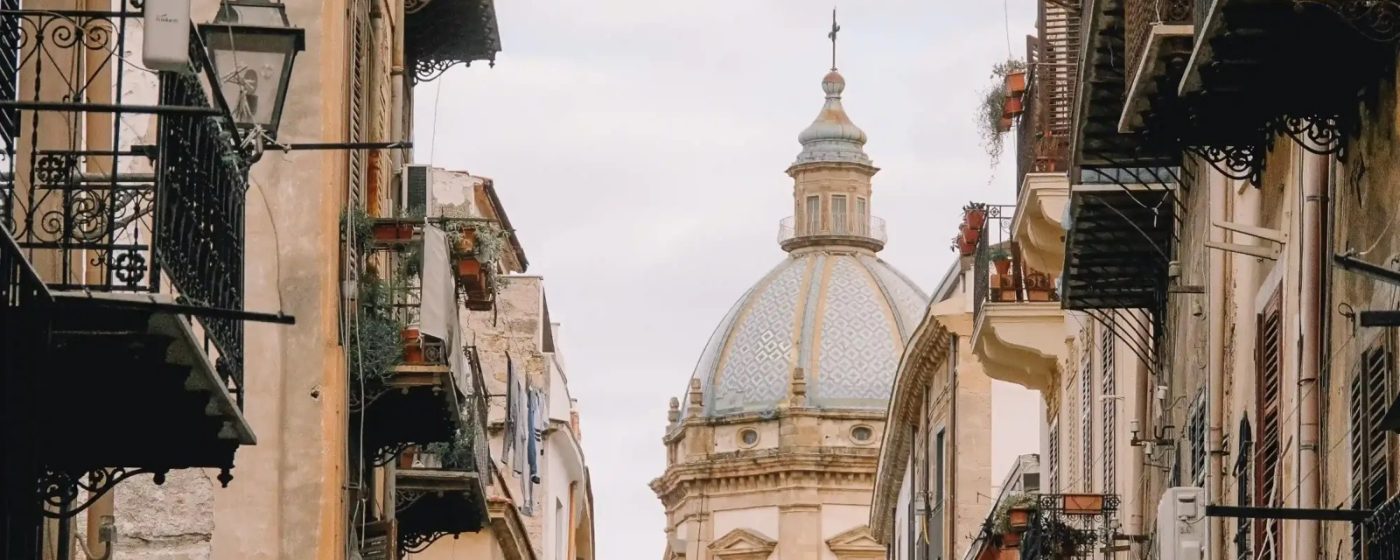 Vicolo del centro storico di Palermo, con balconi in ferro battuto e la cupola maiolicata di una chiesa barocca sullo sfondo